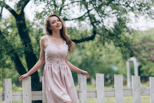 portrait of smiling beautiful woman in stylish dress standing at white fence at countryside