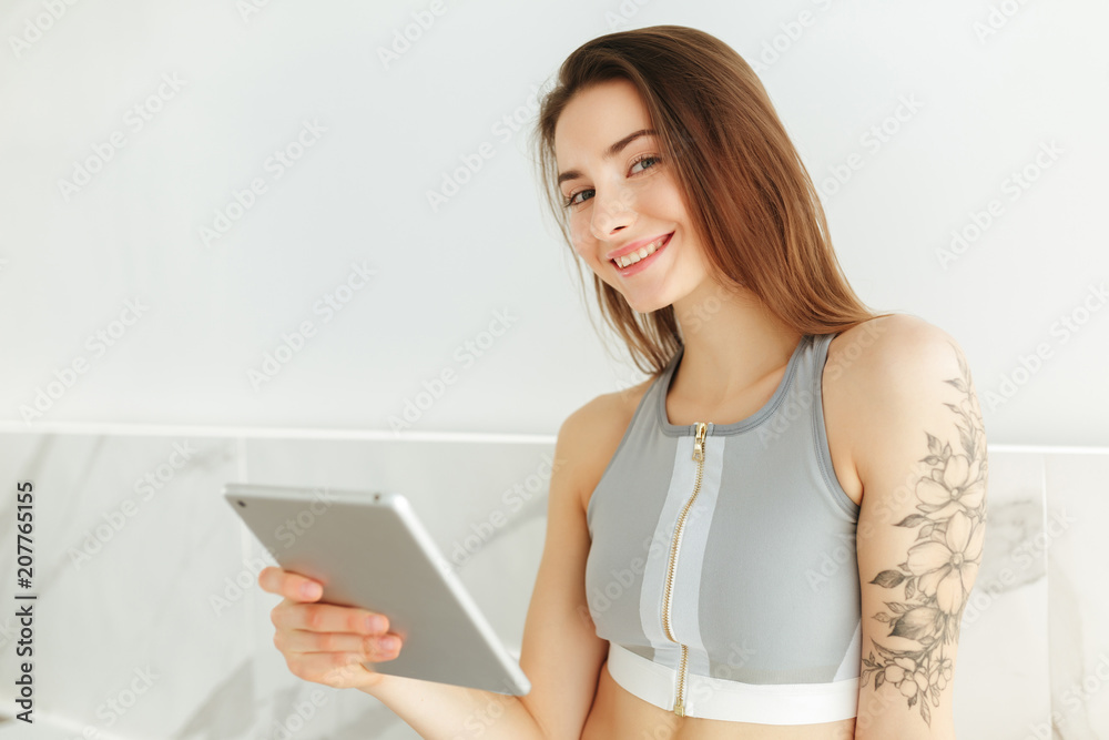 Portrait of young smiling lady in sporty top sitting on kitchen table with tablet in hand and joyfully looking in camera isolated Portrait of young smiling lady in sporty top sitting on kitchen table with tablet in hand and joyfully looking in camera isolated