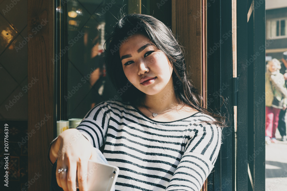 Portrait of a beautiful asian student girl wearing casual outfit smiling at the camera while sitting with a mobile phone in a modern coffee shop. Young blogger having rest in a cafe on a sunny day. Portrait of a beautiful asian student girl wearing casual outfit smiling at the camera while sitting with a mobile phone in a modern coffee shop. Young blogger having rest in a cafe on a sunny day.