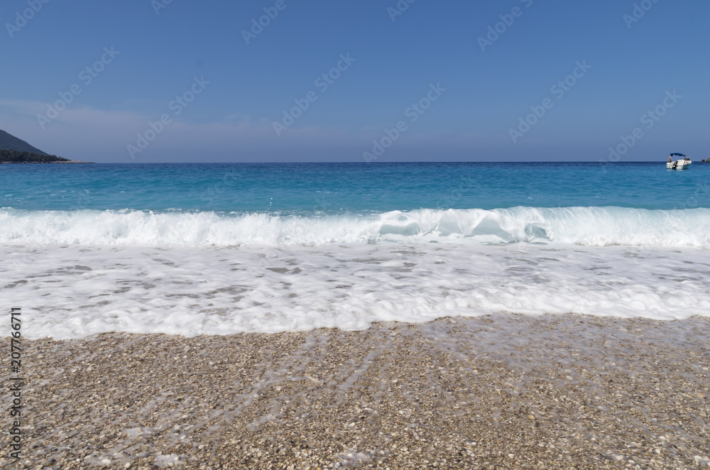 Pebble beach, white sea foam, turquoise sea with a small yacht and blue sky