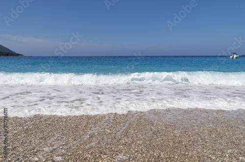 Pebble beach, white sea foam, turquoise sea with a small yacht and blue sky