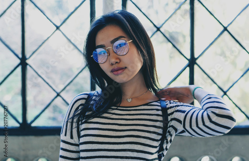 Attractive asian female wearing eyeglasses looking at the camera while standing beside the big window of the spacious hall. Student girl having leisure time at the university campus.