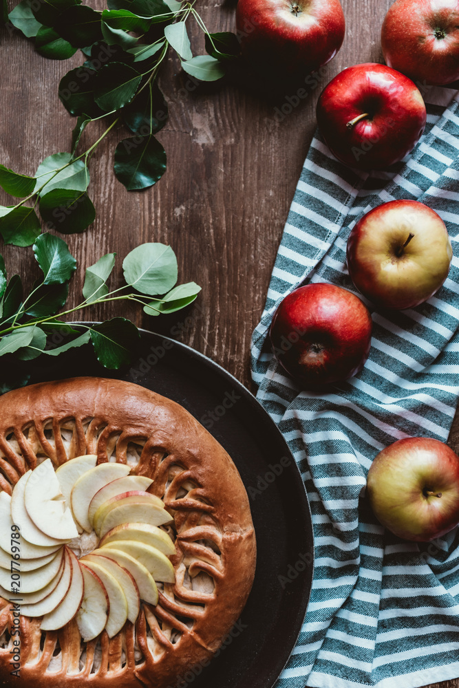 flat lay with arranged homemade apple pie, fresh apples with green leaves and linen on wooden tabletop