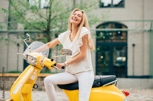 Portrait of sweden smiling blond girl on yellow scooter - Outdoor on street. Retro green modern background. photo