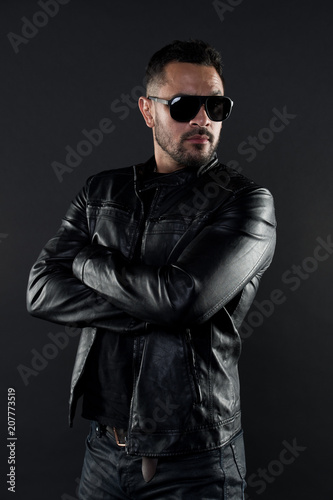 Confident man stand with arms folded. Bearded man in trendy sunglasses. Fashion model in leather jacket and jeans. Fashion and style. Confidence with sexuality and charisma
