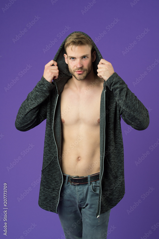 Man on smiling face tries jacket with hood, violet background. Guy with bristle on face and smooth skin on chest tries clothes on. Fashion concept. Man looks attractive in long hooded jacket