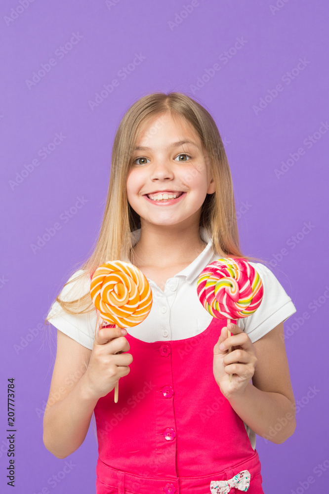 Girl eating big candy on stick or lollipop. Sweet childhood concept. Girl on smiling face holds two giant colorful lollipops in hands, violet background. Kid with long hair likes sweets and treats