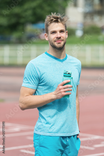 Man with athletic appearance holds bottle with water. Athlete drink water after training at stadium on sunny day. Sport and healthy lifestyle concept. Man athlete in sporty clothes training outdoor