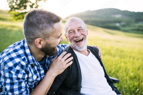 An adult hipster son with senior father in wheelchair on a walk in nature at sunset, laughing. © Halfpoint