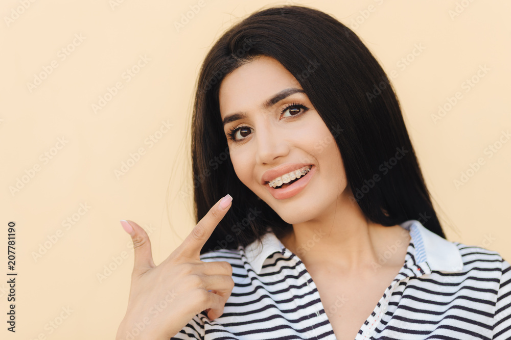 Good looking female with luxurious dark hair, white teeth with brackets, indicates at mouth with fore finger, advertises braces, being delighted after visit to dentist, isolated over studio background Good looking female with luxurious dark hair, white teeth with brackets, indicates at mouth with fore finger, advertises braces, being delighted after visit to dentist, isolated over studio background
