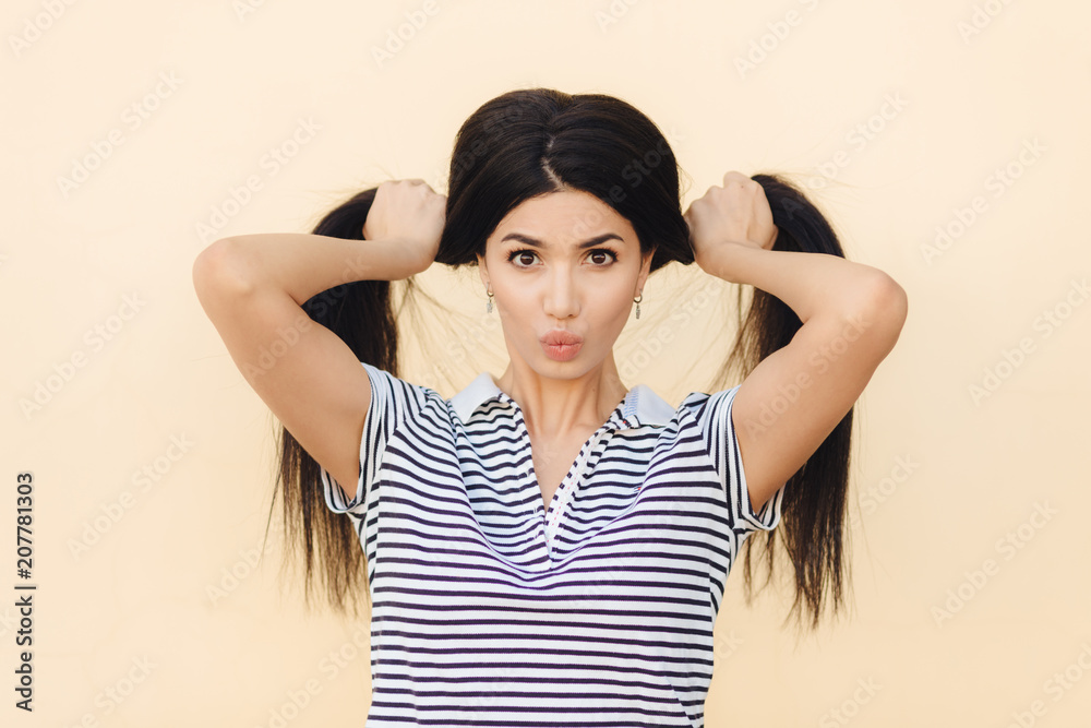 Portrait of pretty brunette female model makes two pony tails from her luxurious hair, has dark eyes, keeps lips round, wears casual t shirt, isolated over light studio background. People and fashion Portrait of pretty brunette female model makes two pony tails from her luxurious hair, has dark eyes, keeps lips round, wears casual t shirt, isolated over light studio background. People and fashion