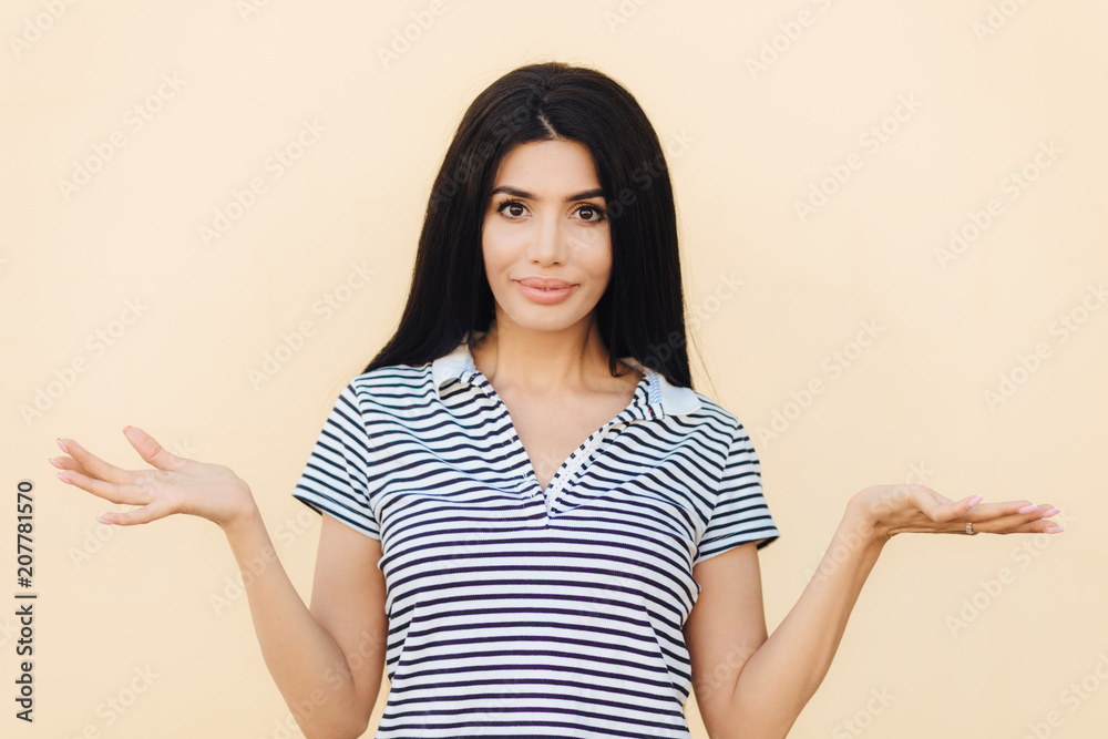 Who cares? Adorable brunette young female with hesitant expression, shrugs shoulders in bewilderment, tries to find solution or makes decision, poses indoor alone. People and hesitation concept Who cares? Adorable brunette young female with hesitant expression, shrugs shoulders in bewilderment, tries to find solution or makes decision, poses indoor alone. People and hesitation concept