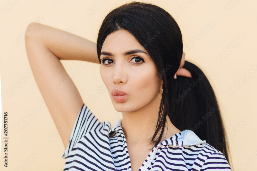 Lovely female with dark hair, make up, pouts lips, makes pony tail, has healthy skin, dressed in casual clothes, isolated over light background. People, beauty and facial expressions concept Lovely female with dark hair, make up, pouts lips, makes pony tail, has healthy skin, dressed in casual clothes, isolated over light background. People, beauty and facial expressions concept