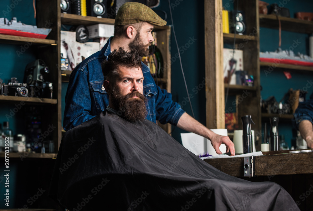 Man with beard covered with black cape waiting while barber changing clipper grade. Hipster client getting haircut. Client with beard ready for trimming or grooming. Haircut process concept Man with beard covered with black cape waiting while barber changing clipper grade. Hipster client getting haircut. Client with beard ready for trimming or grooming. Haircut process concept
