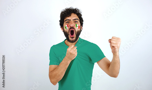 Sport fan screaming for the triumph of his team. Man with the flag of Mexico makeup on his face and green t-shirt.