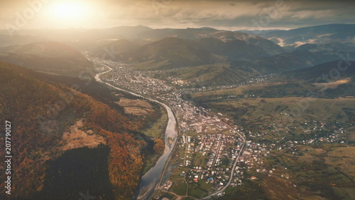 Aerial view over dramatic autumn sunset landscape. Small mountain village in canyon surrounded meadows, orange hills, pine tree forests. Carpathians, Ukraine, Europe. Vintage retro dark toning filter.