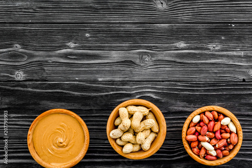 Peanut paste concept. Bowls with butter, nuts in shell, peeled nuts on black wooden background top view copy space