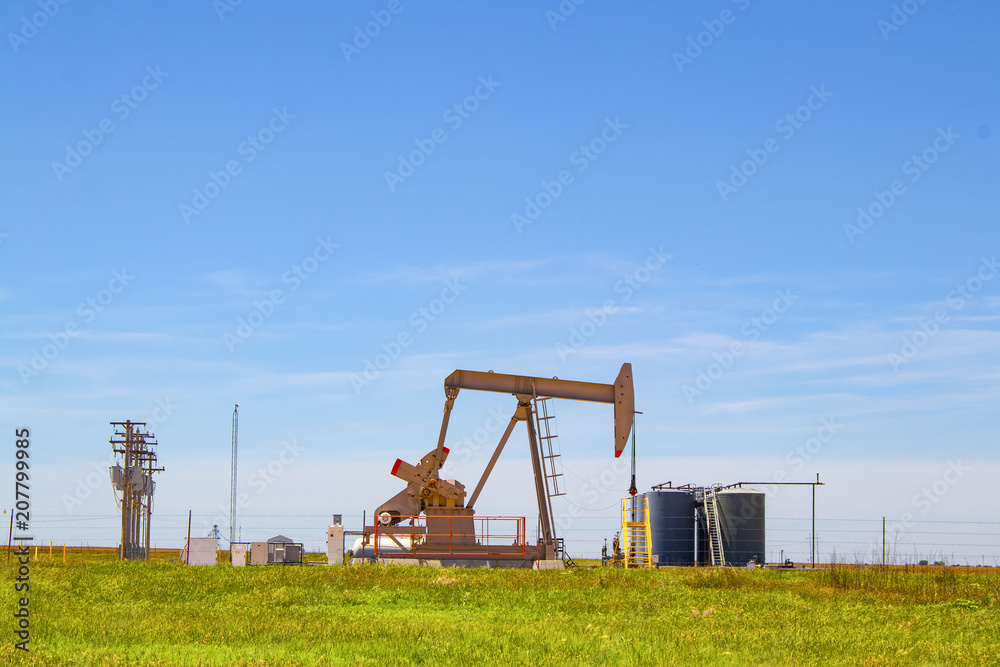 Working Pump Jack on oil well with  tanks on site out on the horizon on the plains with electric lines and blue sky in background