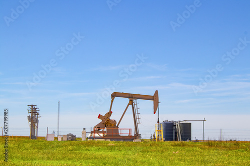 Working Pump Jack on oil well with tanks on site out on the horizon on the plains with electric lines and blue sky in background