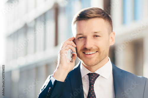 Outdoor shot of prosperous successful unshaven businessman wears formal clothes, has pleasant talk with business partner via cell phone, discuss future meeting, poses against blurred background photo