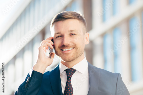 Glad handsome cheerful male entrepreneur wears formal suit, tie and white shirt, holds modern cell phone, communicates with business partner, discusses contract terms. People and occupation. photo