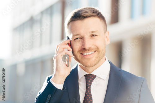 Portrait of delightful European businessman, wears elegant suit, talks on smart phone, deals with business partner, decide time of meeting, has positive expression. People, career, technology concept photo