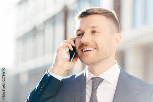 Portrait of successful positive office employee has phone conversation, looks happily into distance, discusses money payment. Male executive manager in formal suit speaks with client via cellular photo