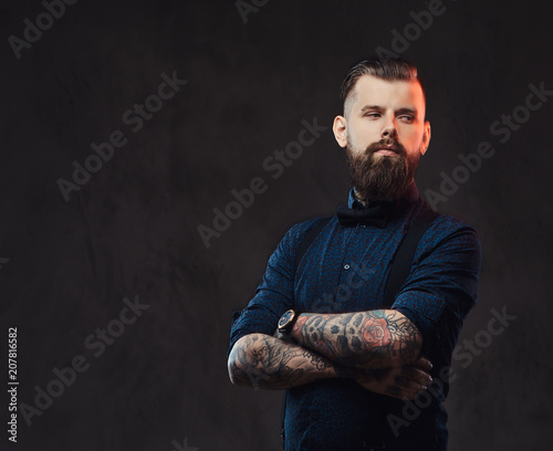 Portrait of a handsome old-fashioned hipster in a blue shirt and suspenders, standing with crossed arms in a studio. © Fxquadro