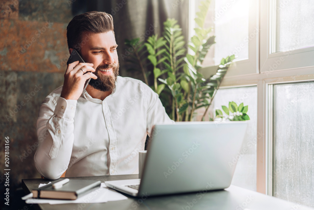 Young smiling bearded businessman is sitting at table in front of laptop, drinking coffee, talking on cell phone. Telephone conversations, distance work, online marketing, education, e-commerce. Young smiling bearded businessman is sitting at table in front of laptop, drinking coffee, talking on cell phone. Telephone conversations, distance work, online marketing, education, e-commerce.