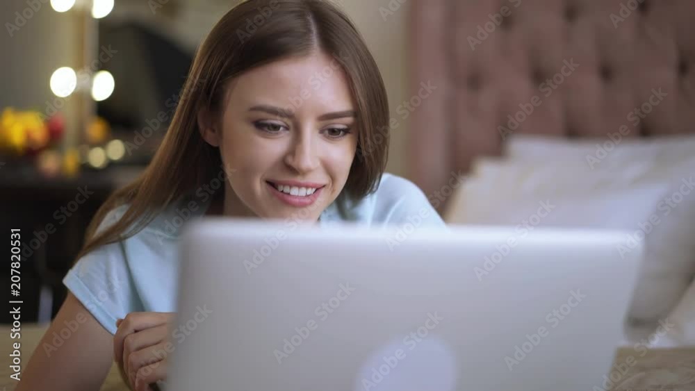 Portrait of a young woman with long fair hair lying on her bed and web surfing on her laptop computer. Left to right pan real time medium shot