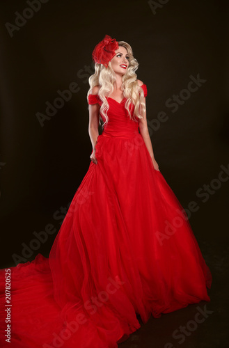 Blonde woman in red dress and elegant hat isolated on studio black background. Beautiful blond girk with long wavy hair posing in prom gown.