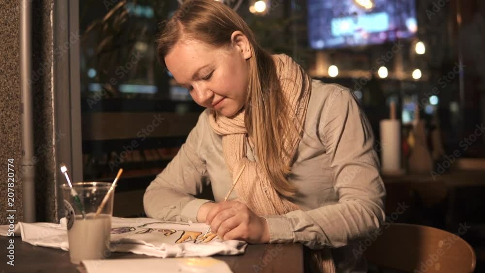 Skilled young woman is drawing beautiful figure on a fabric of white clothing. She is sitting in cafe in evening time alone Skilled young woman is drawing beautiful figure on a fabric of white clothing. She is sitting in cafe in evening time alone