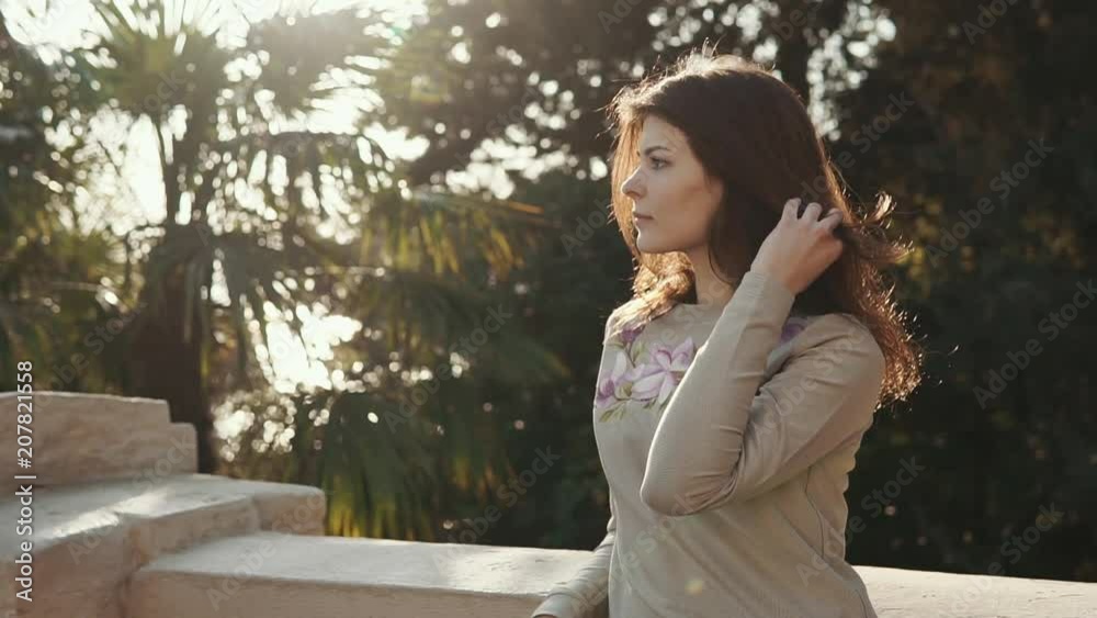 Pretty brunette girl is touching her hair, standing in sunny terrace of garden. She is looking on side and breathing fresh morning air Pretty brunette girl is touching her hair, standing in sunny terrace of garden. She is looking on side and breathing fresh morning air