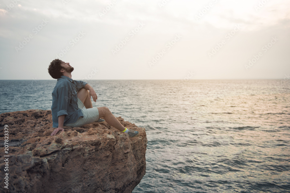 Full length profile of delighted young guy with beard is sitting on the edge of cliff while raising head up to sky with closed eyes. He is dreaming about his future. Copy space in the right side Full length profile of delighted young guy with beard is sitting on the edge of cliff while raising head up to sky with closed eyes. He is dreaming about his future. Copy space in the right side