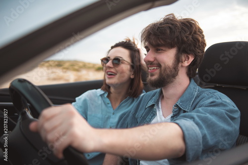 Summertime. Cheerful young pleasant male and female are travelling by luxury car. Focus on cute bearded boyfriend is holding on to steering wheel and expressing gladness. Romantic trip concept