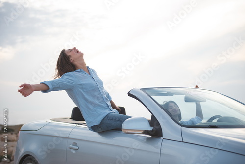 Full of admiration. Happy girl is sitting on fashionable cabriolet with her hands outstretched while expressing rapture. She is travelling by car along seashore with her friend female. Summer concept