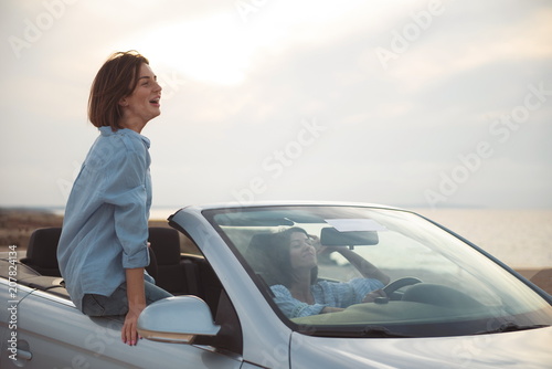 Happy vacation. Positive young women are travelling by fashionable cabriolet with open roof. They are enjoying their summer holiday abroad. Girl is sitting on car and looking ahead with smile