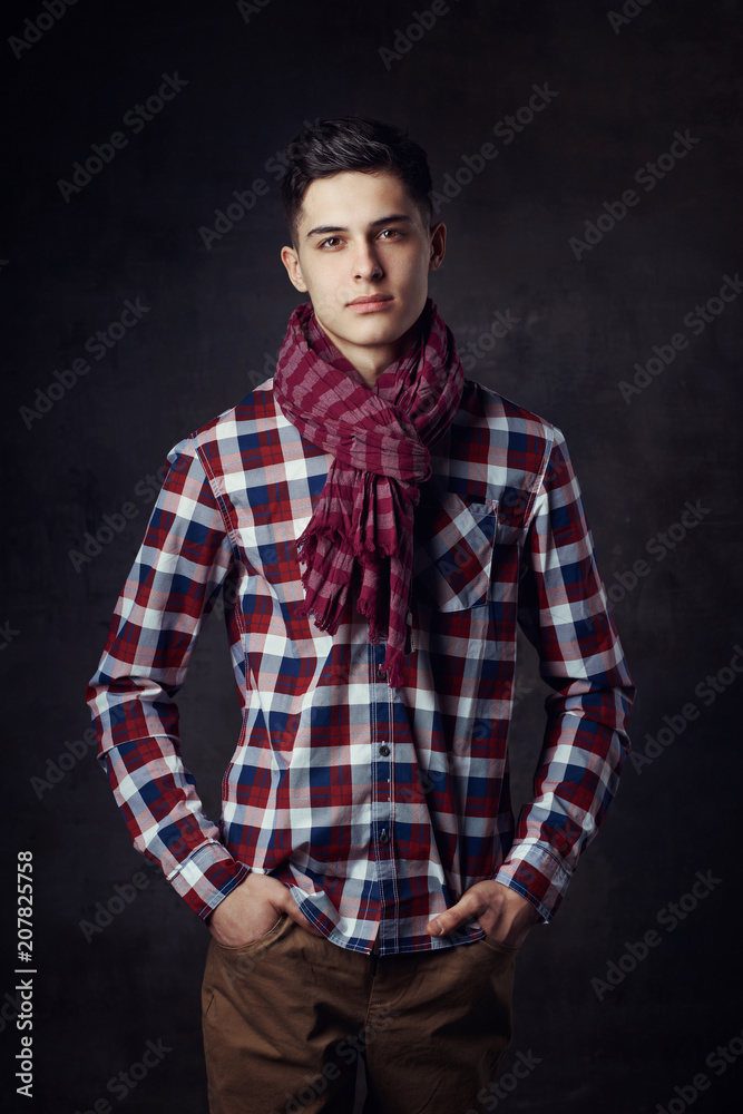 Portrait of young trendy handsome man with short dark hair wearing checkered shirt and brown trousers standing and posing against gray concrete wall Portrait of young trendy handsome man with short dark hair wearing checkered shirt and brown trousers standing and posing against gray concrete wall