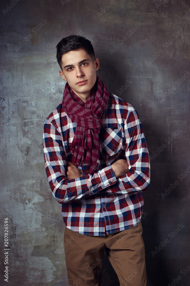 Portrait of young trendy handsome man with short dark hair wearing checkered shirt and brown trousers standing and posing against gray concrete wall Portrait of young trendy handsome man with short dark hair wearing checkered shirt and brown trousers standing and posing against gray concrete wall