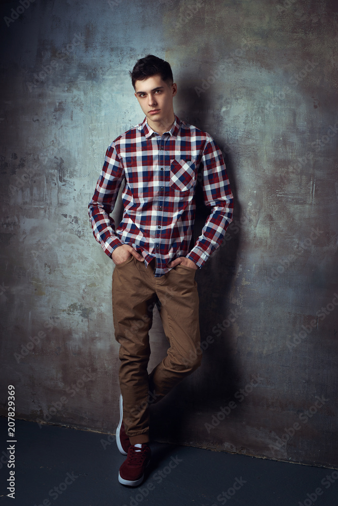 Portrait of young trendy handsome man with short dark hair wearing checkered shirt and brown trousers standing and posing against gray concrete wall Portrait of young trendy handsome man with short dark hair wearing checkered shirt and brown trousers standing and posing against gray concrete wall