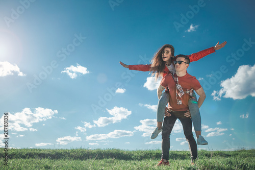 I believe we can fly. Full length of amorous couple looking at blue sky. Girl is on her boyfriend back with stretched arms. They smiling and enjoying summer weather. Copy space in left side