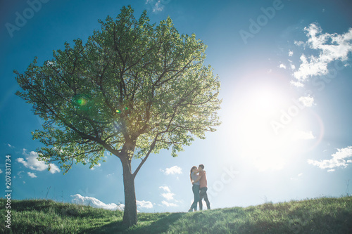 Love is in the air. Full length of nice loving people embracing on hill by tree. They are giving kisses to each other with exciting summer landscape on background
