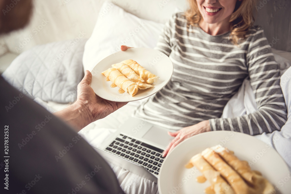 Top view focus on white plate with sweet rolls with topping. Gentle man is giving breakfast to soulmate while she is sitting in bed with laptop. Grateful woman is taking dish and smiling with delight Top view focus on white plate with sweet rolls with topping. Gentle man is giving breakfast to soulmate while she is sitting in bed with laptop. Grateful woman is taking dish and smiling with delight