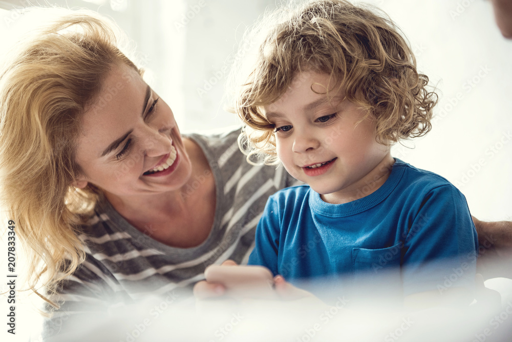 Cheerful mom is enjoying time with little boy while he is using mobile phone. Cute kid is attentively handling mobile phone with great interest. Children and modern gadgets concept Cheerful mom is enjoying time with little boy while he is using mobile phone. Cute kid is attentively handling mobile phone with great interest. Children and modern gadgets concept
