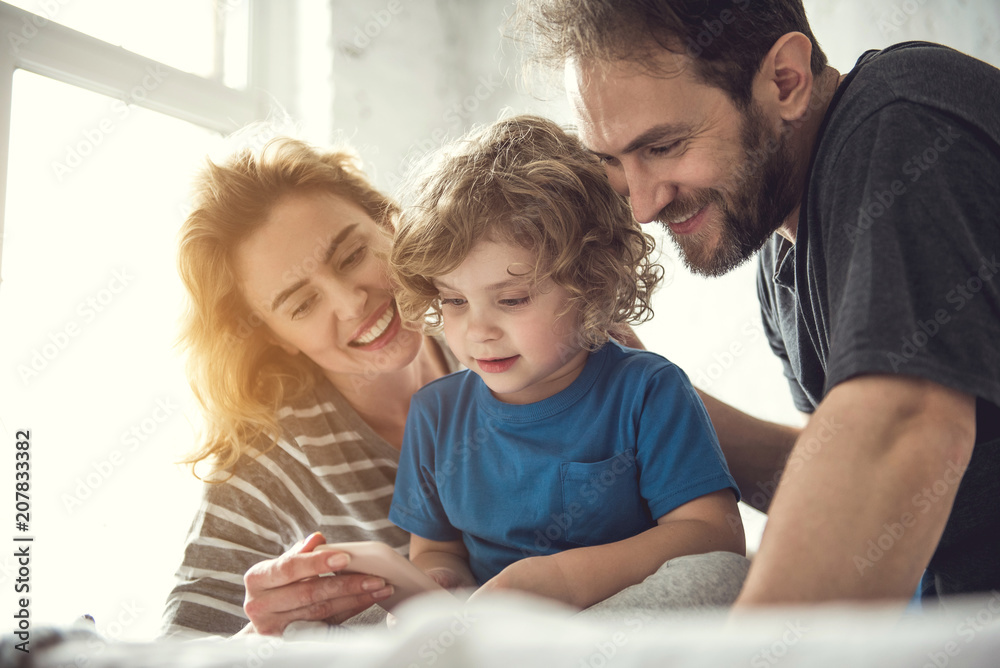 Loving spouse is showing smartphone to their exited son. Father is tenderly touching boy on back while mom is holding gadget. All family are spending time with modern technologies concept Loving spouse is showing smartphone to their exited son. Father is tenderly touching boy on back while mom is holding gadget. All family are spending time with modern technologies concept