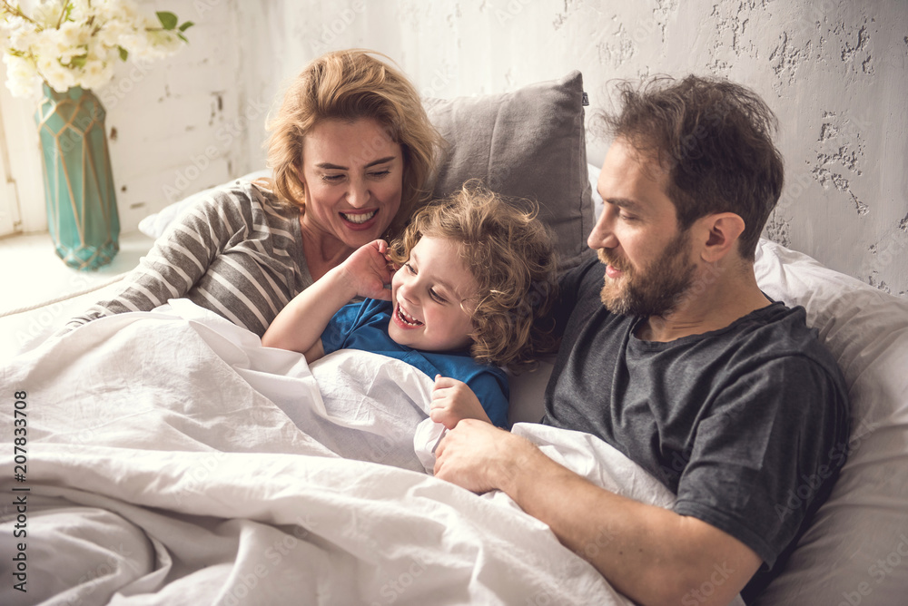 Obraz premium Carefree spouse is cuddling pretty son in large comfortable bed. They are sharing happy emotions with each other as day light is coming into cozy apartment