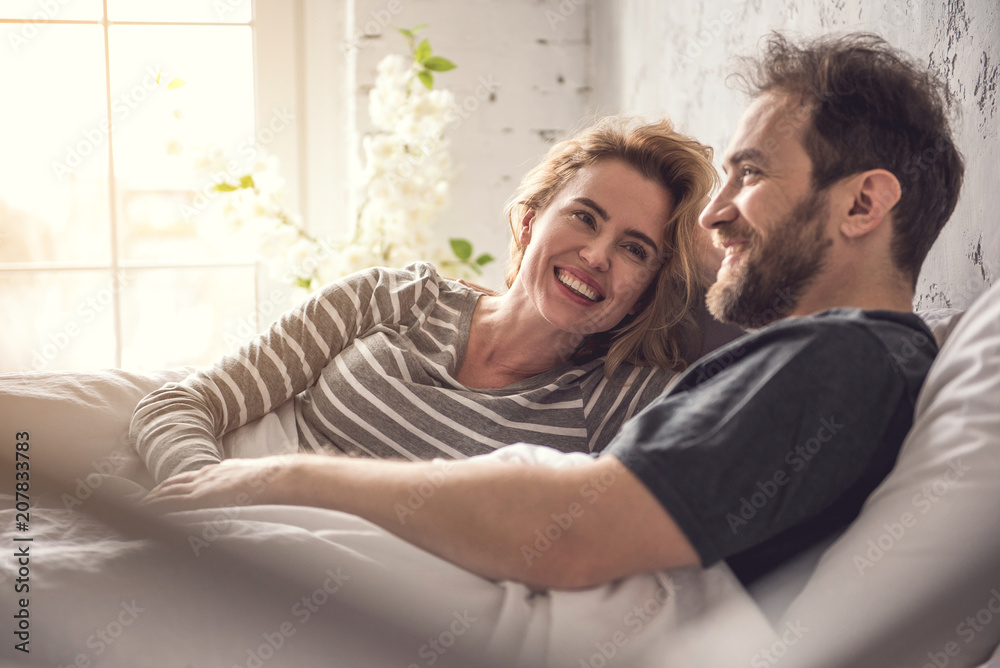 Attractive lady is smiling to partner while they are hanging out in dormitory during morning time. Relaxed carefree man is looking ahead and gently taking woman hand Attractive lady is smiling to partner while they are hanging out in dormitory during morning time. Relaxed carefree man is looking ahead and gently taking woman hand