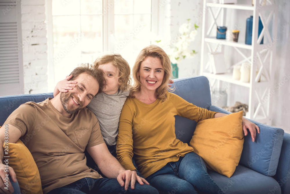 Cheerful parents are sitting with son in nice sitting-room and bonding child. Kid is kissing his doting father while man is closing eyes with bliss. Calm grinning mother is watching at camera