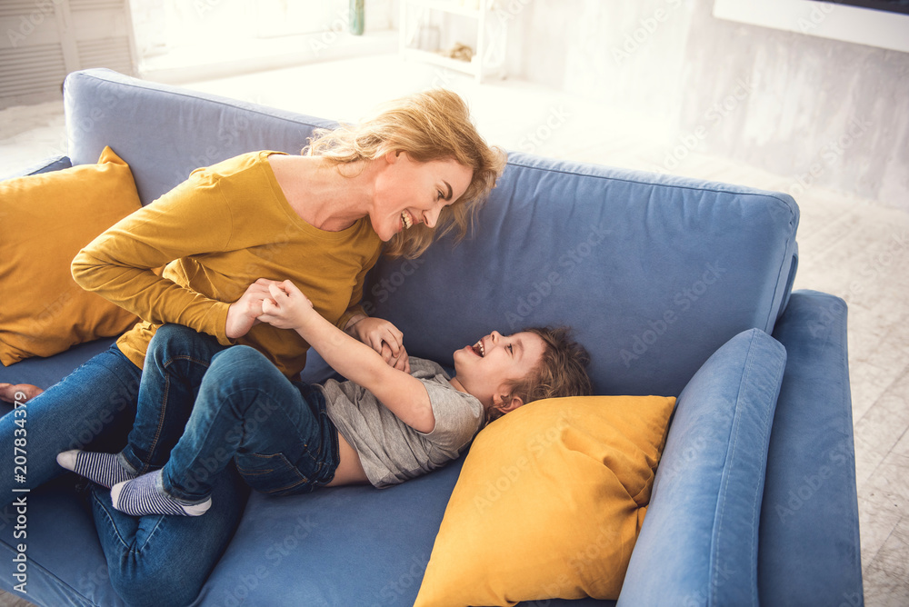 Top view of happy family playing together on couch indoors. Laughing mother and kid are hand-holding and looking into each-other eyes. Parents are playing with children at home concept