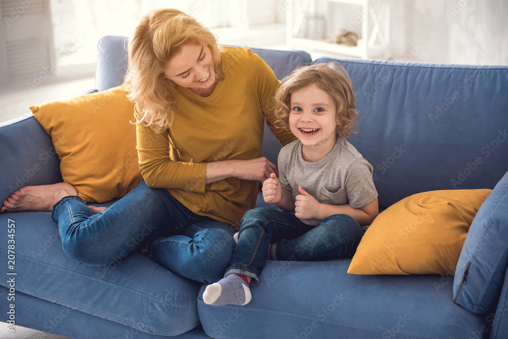 Happy family are having fun together in their comfortable house. They are sitting on sofa while mother is lovingly touching little boy. Cute child is looking at camera with joy